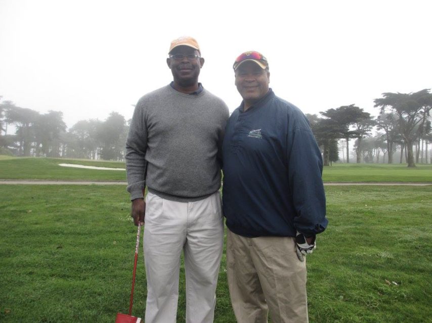 Two men standing together on a golf course, one holding a golf club, with trees and fog in the background.