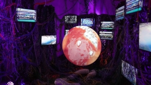 A glowing sphere resembling a planet is surrounded by multiple screens displaying static and data, with tangled wires in a dimly lit, purple room.