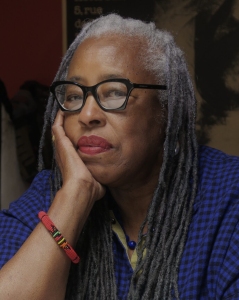 An older woman with long gray dreadlocks, glasses, and a thoughtful expression rests her face on her hand. She wears a blue checkered shirt and a red bracelet.