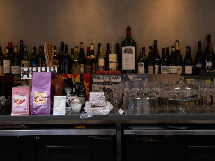 Bottles of wine, glasses, coffee bags, and packaged goods are arranged on a bar counter against a gray wall.