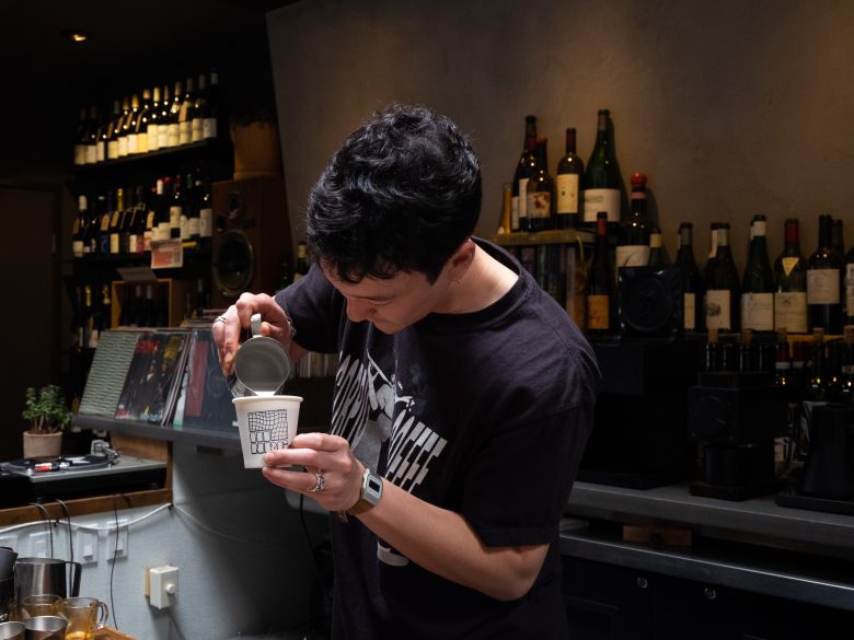 A person pours milk from a metal pitcher into a paper cup at a bar or café with shelves of wine bottles in the background.
