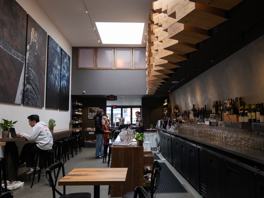 Modern bar with high ceiling, geometric wooden decor, wall art, a long counter with bottles and glasses, and a few people seated or standing inside.