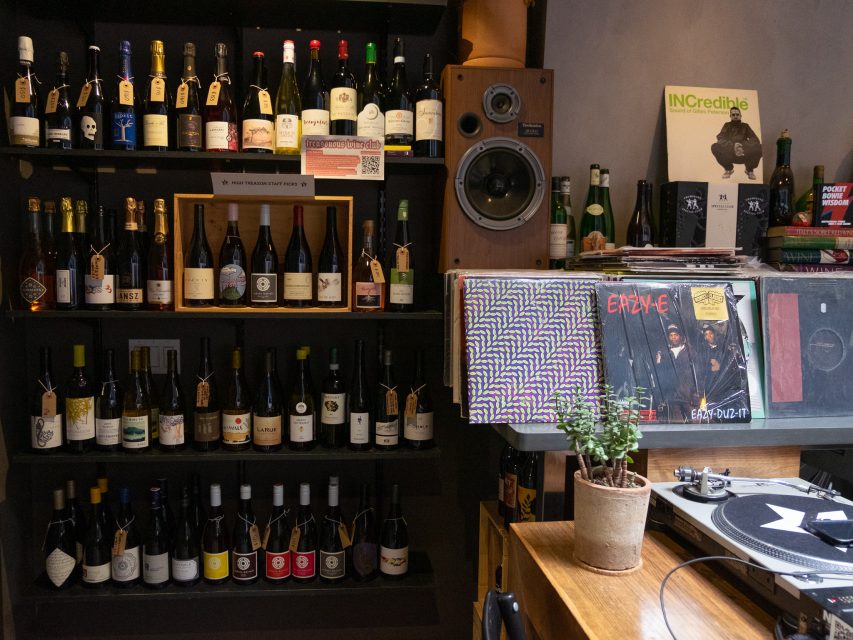 Shelves filled with wine bottles and a few beer bottles, a speaker, vinyl records including N.W.A and Eazy-E, a potted plant, and a turntable on a wooden surface.