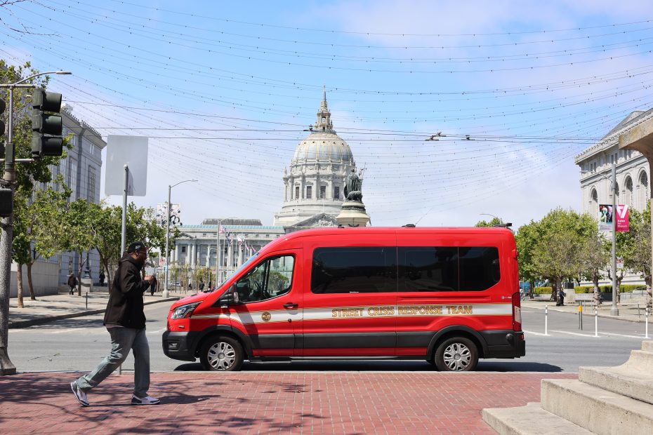 A red van labeled "Street Crisis Response Team" is parked on a city street near a domed government building; in the foreground, a person walks by, echoing the urban scenes often captured by Joanna Sokol.