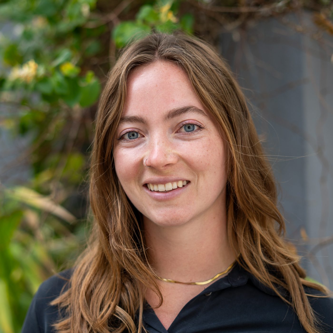 A woman with long light brown hair and blue eyes smiles at the camera, wearing a black top and gold necklace, with greenery in the background.