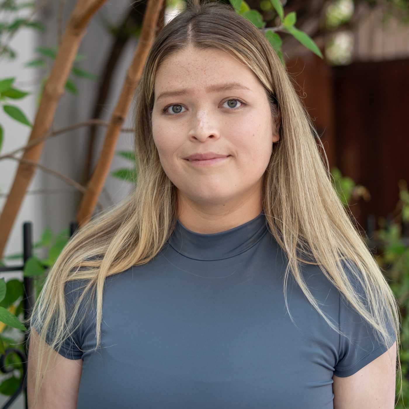 A woman with long blonde hair wearing a gray top stands outdoors in front of greenery, looking at the camera with a neutral expression.