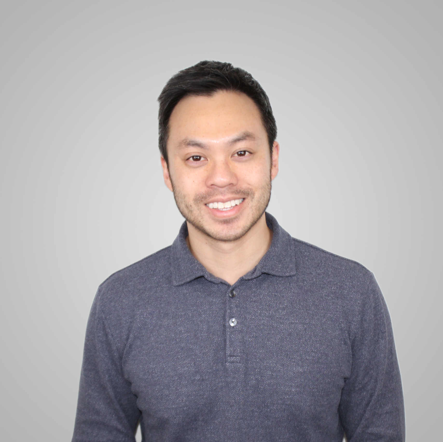 A man with short dark hair and a trimmed beard, wearing a dark grey polo shirt, stands facing the camera and smiles against a plain light grey background.