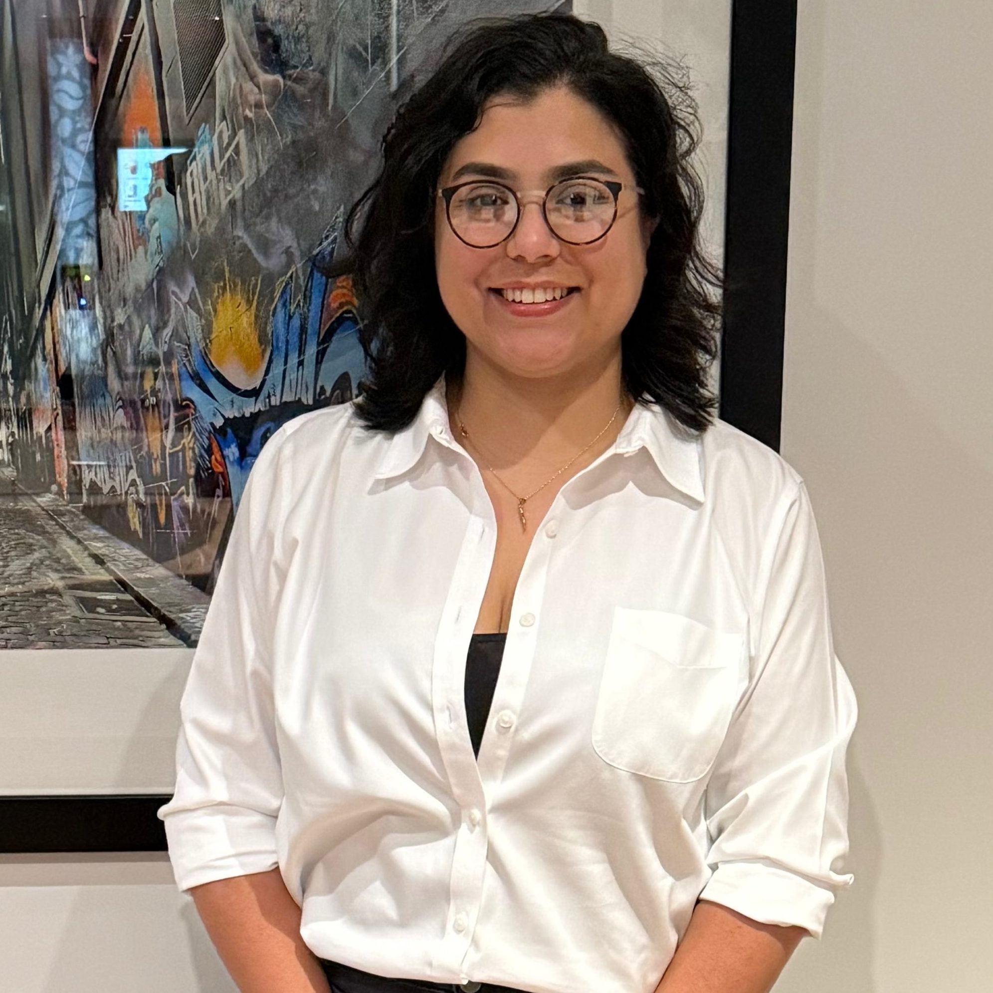 A person with dark hair and glasses smiles while wearing a white shirt, standing in front of a framed artwork featuring urban graffiti.