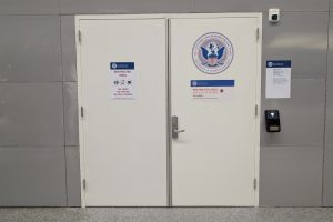 White double doors labeled "Restricted Area" with a U.S. Department of Homeland Security seal, access keypad, and security notices on a gray wall.