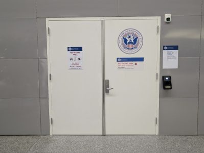 White double doors labeled "Restricted Area" with a U.S. Department of Homeland Security seal, access keypad, and security notices on a gray wall.