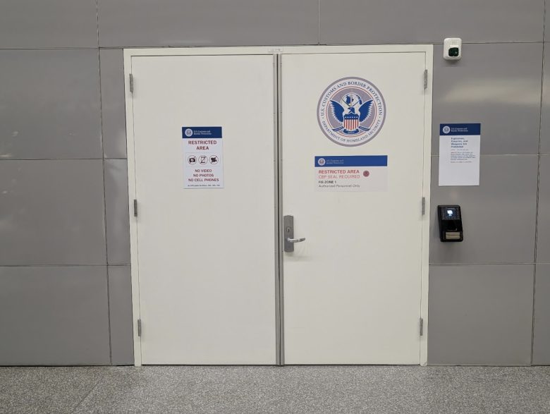 White double doors labeled "Restricted Area" with a U.S. Department of Homeland Security seal, access keypad, and security notices on a gray wall.
