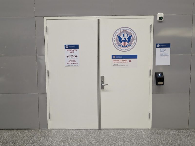 White double doors labeled "Restricted Area" with a U.S. Department of Homeland Security seal, access keypad, and security notices on a gray wall.