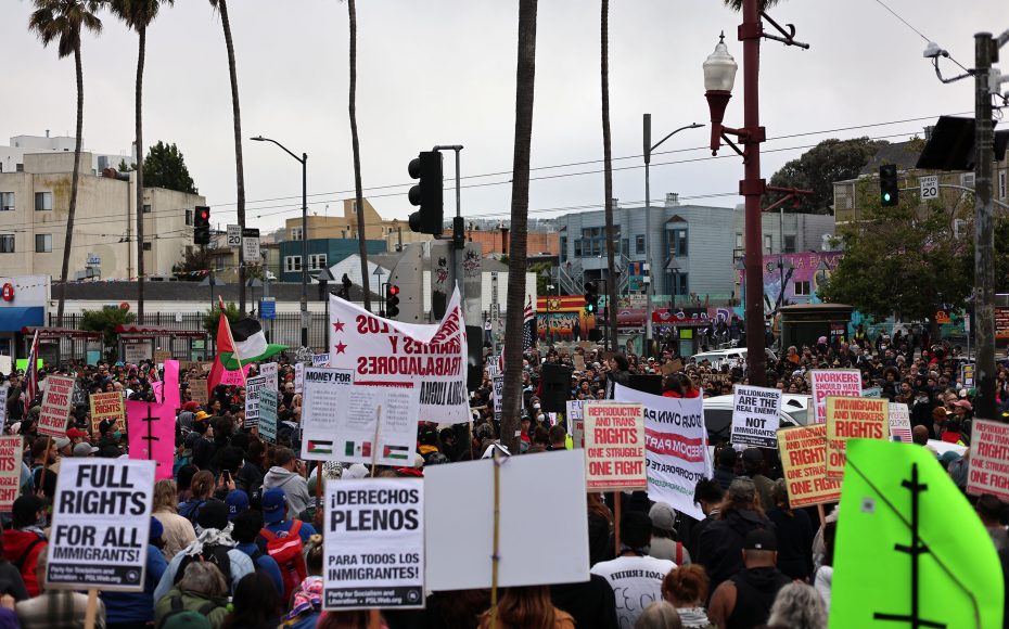 Large crowd at a street protest holds signs demanding full rights for immigrants and workers, with some calling to abolish ICE; buildings and palm trees line the background.