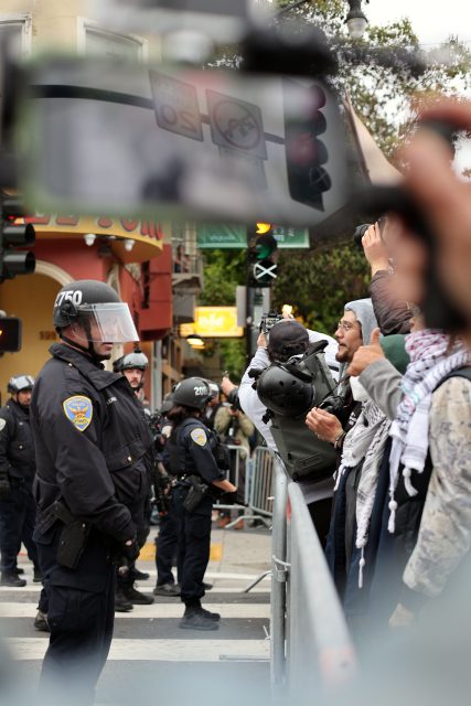 Police officers in riot gear and ICE agents stand facing a group of protesters behind a barricade on a city street during a daytime demonstration.