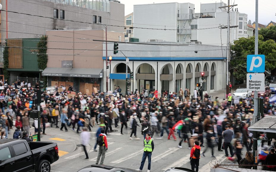 A large crowd of people gathers and marches at a city intersection while two uniformed personnel, possibly discussing ICE enforcement, direct traffic nearby.