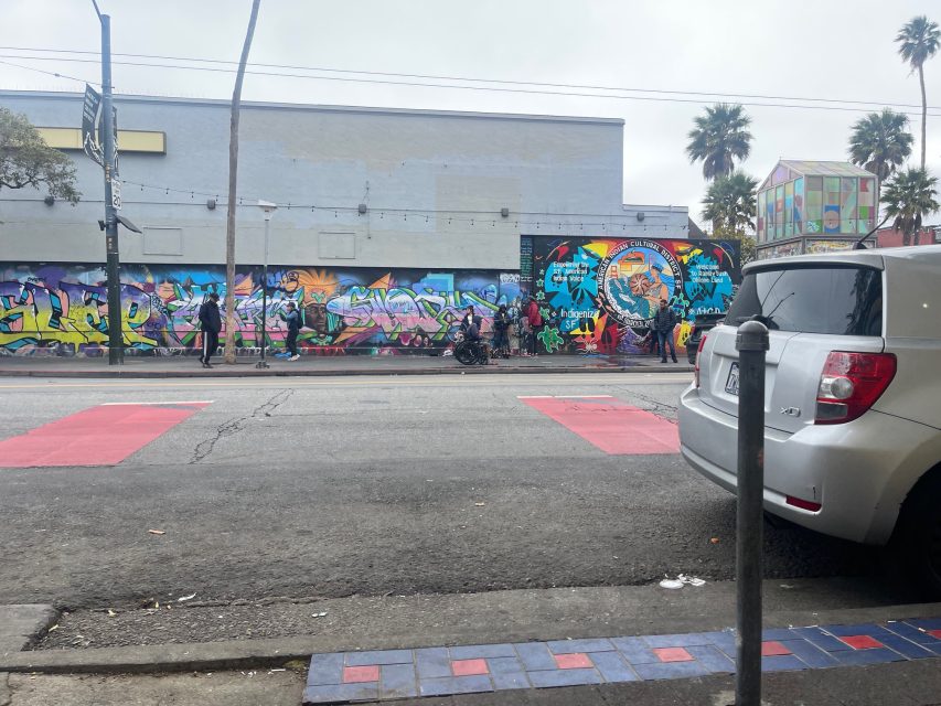 Street view of a colorful graffiti mural on a long wall with several pedestrians walking by; parked car and tiled sidewalk in the foreground.