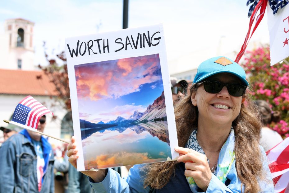 A woman wearing sunglasses and a blue cap holds a sign with a landscape photo labeled "WORTH SAVING" at an outdoor protest. An American flag is visible in the background.