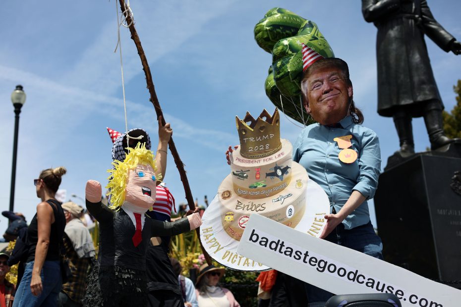 A person wearing a Trump mask holds a decorated cake prop and a banner reading "badtastegoodcause.com" at an outdoor protest, with others and a Trump piñata nearby.