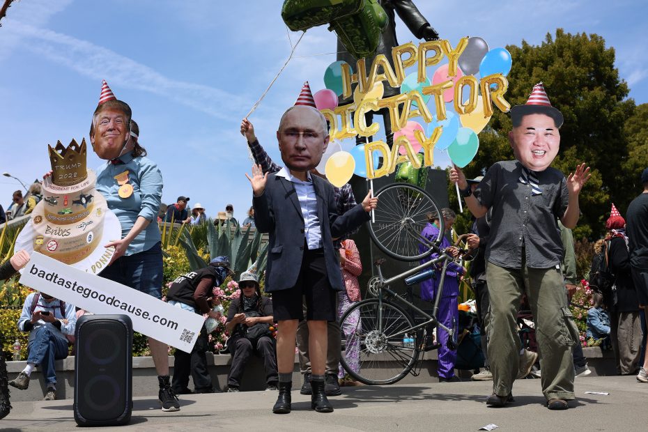 Three people wearing oversized masks and party hats depicting world leaders pose with a "Happy Dictator Day" sign during a public outdoor protest.
