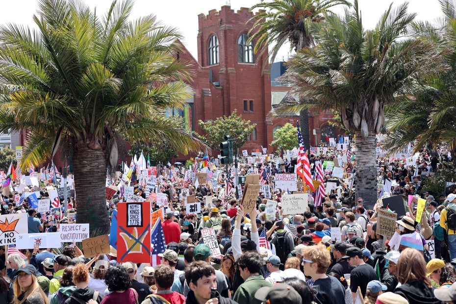 A large protest draws a crowd outdoors holding signs, with palm trees and a red brick building visible in the background.