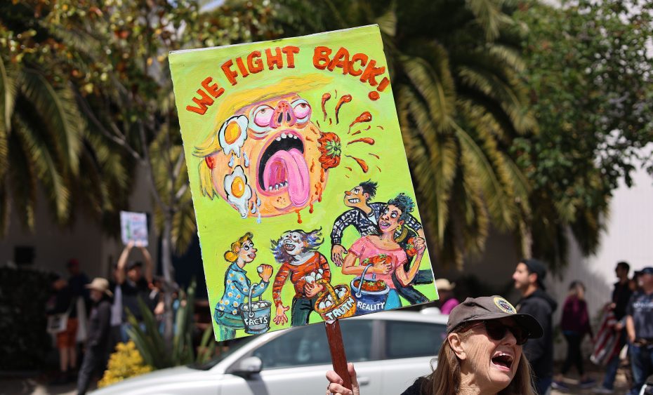 A protester holds a colorful sign reading "WE FIGHT BACK!" with cartoonish figures; a protest unfolds as people march in the background near palm trees.