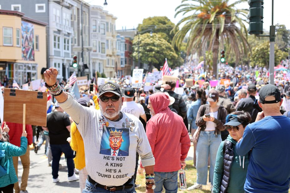 At a protest, a man raises his fist. He wears sunglasses, hats, pins, and a shirt with a caricature of Donald Trump and the text, “He’s not a king, he’s a dumb loser,” as the crowd gathers behind him.