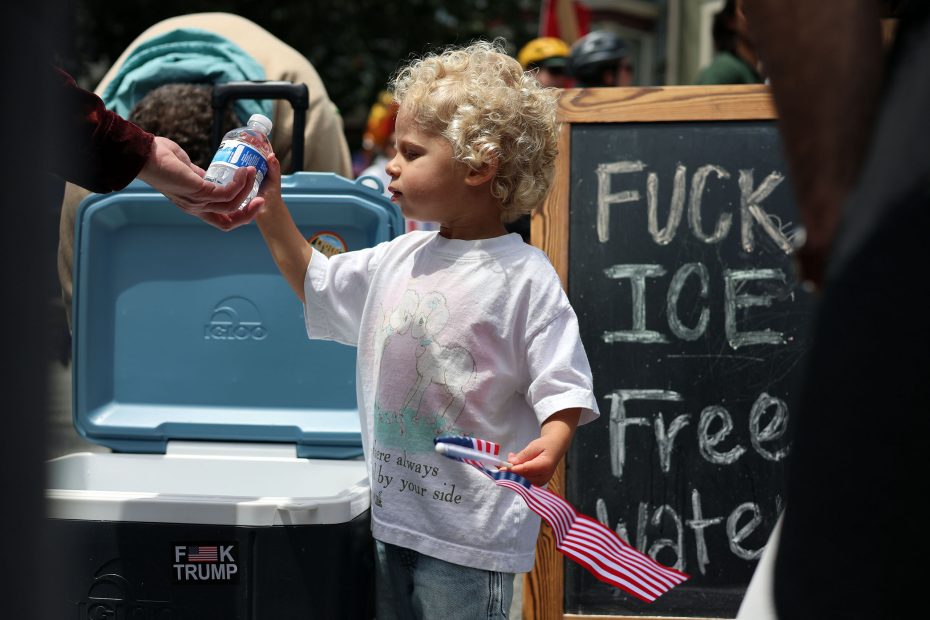 A young child receives a water bottle near a cooler, standing by a chalkboard sign at a protest offering free water with a profane message about ICE.