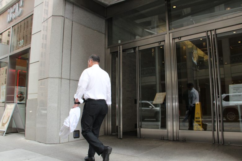 A man in a white shirt and black pants walks toward a glass entrance of a modern office building, holding a jacket.
