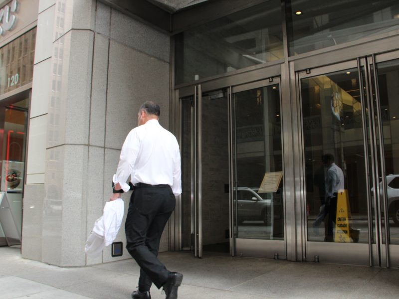 A man in a white shirt and black pants walks toward a glass entrance of a modern office building, holding a jacket.