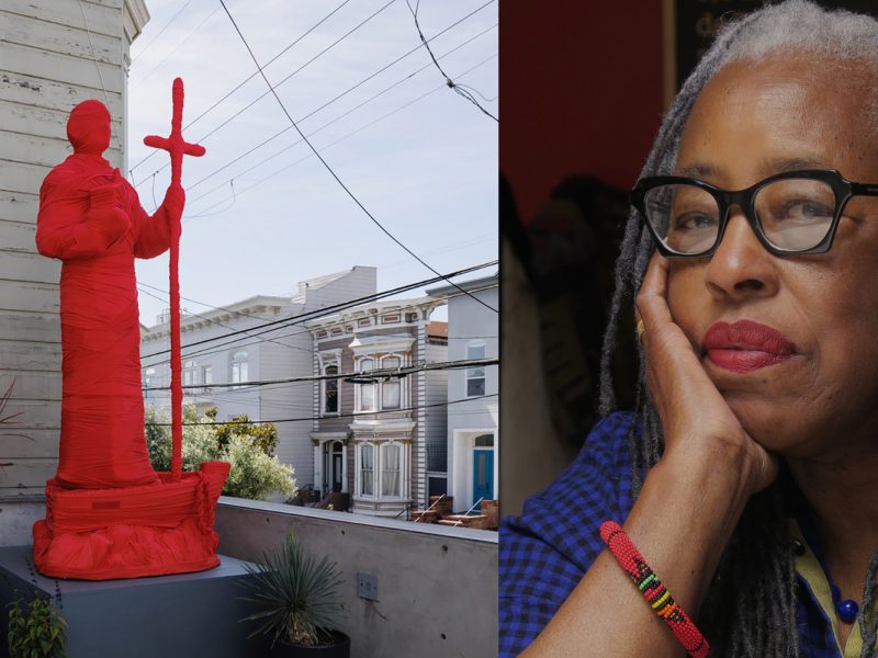 A red statue of a standing figure holding a cross is outdoors; next to it, an older woman with gray hair and glasses rests her chin on her hand, looking thoughtful.