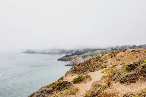 A coastal landscape with rugged cliffs, sparse vegetation, and a dirt path overlooking the ocean under a foggy sky.