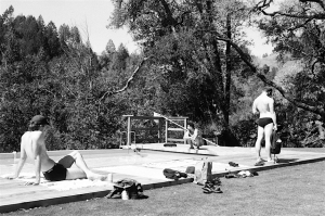 Three people in swimsuits relax near a wooden dock by a body of water, surrounded by trees and scattered belongings, on a sunny day.