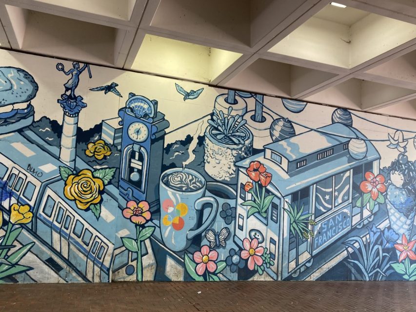 A mural depicts San Francisco icons including a cable car, flowers, a coffee cup, and a clock tower, all painted in blue tones with pops of color.