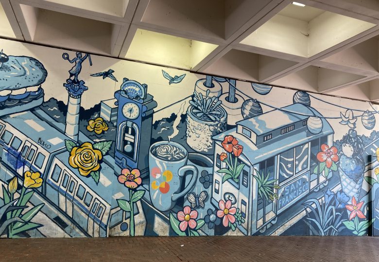A blue-toned mural features a San Francisco cable car, a clock tower, flowers, a coffee cup, a burger, and birds in a whimsical cityscape.