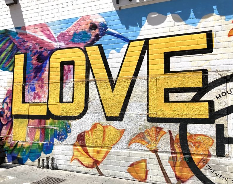 A colorful mural on a white brick wall features a large yellow "LOVE" with a vibrant hummingbird and yellow flowers surrounding the letters.