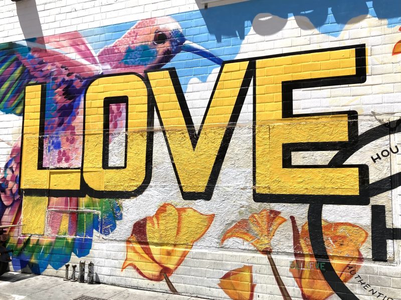 A colorful mural on a white brick wall features a large yellow "LOVE" with a vibrant hummingbird and yellow flowers surrounding the letters.
