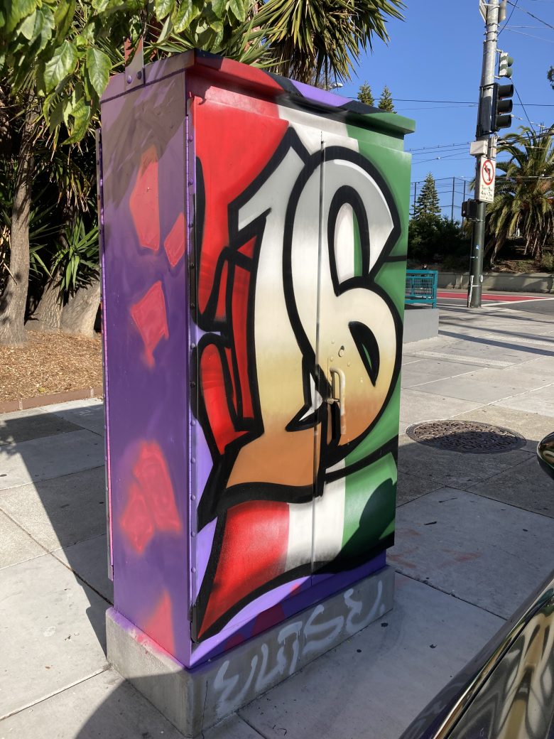A utility box on a sidewalk painted with large graffiti-style numbers "16" over a red, white, and green background, with purple sides and nearby street signs visible.