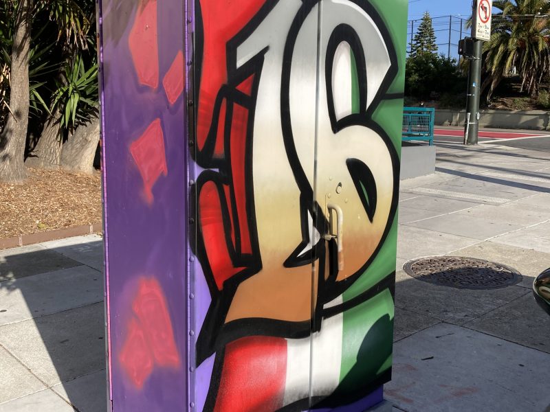 A utility box on a sidewalk painted with large graffiti-style numbers "16" over a red, white, and green background, with purple sides and nearby street signs visible.