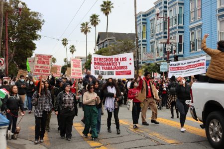 A diverse group of people march on a city street holding signs advocating for immigrant and workers' rights.