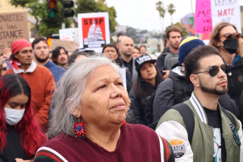 A diverse group of people, some holding protest signs, gathers outdoors. A woman with gray hair and beaded earrings stands in the foreground, looking ahead.