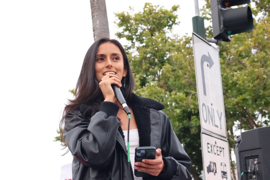 A woman wearing a black jacket speaks into a microphone at an outdoor event, holding a phone. A traffic sign and trees are visible in the background.