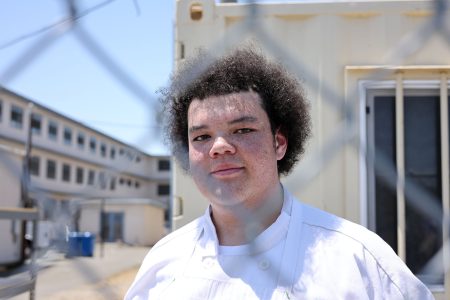A person wearing a white chef's coat stands outside near a chain-link fence, with buildings and a blue sky in the background.