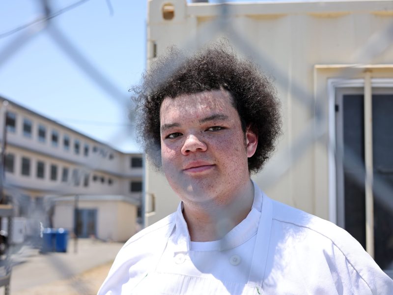 A person wearing a white chef's coat stands outside near a chain-link fence, with buildings and a blue sky in the background.