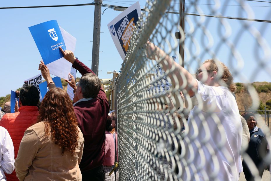 A group of people hold signs and papers while standing behind a chain-link fence during a daytime outdoor gathering.