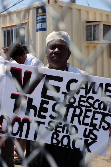 A person stands behind a fence holding a sign that reads, "WE CAME TO ESCAPE THE STREETS. DON'T SEND US BACK." Several other people are visible in the background.