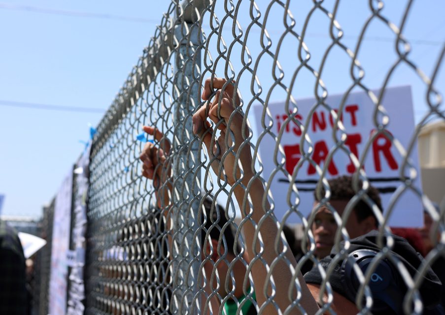 Hands gripping a chain-link fence, with people holding protest signs visible in the background.
