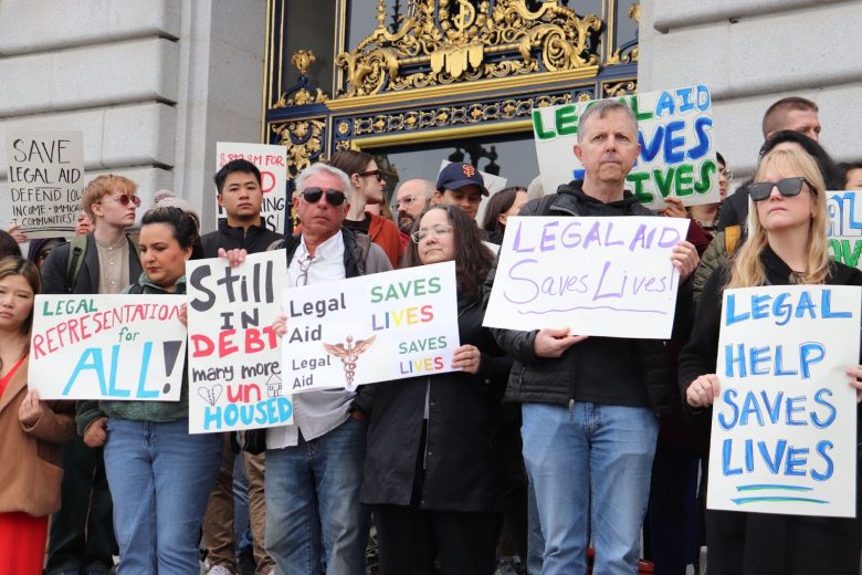 A group of people stand outside a building holding signs advocating for legal aid, with messages such as “Legal Aid Saves Lives” and “Legal Representation for All.”.