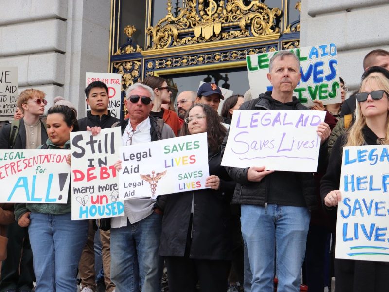 A group of people stand outside a building holding signs advocating for legal aid, with messages such as “Legal Aid Saves Lives” and “Legal Representation for All.”.