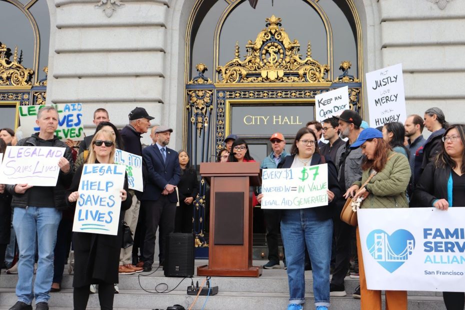 A group of people gather on city hall steps holding signs supporting legal aid and social justice; a podium is set up in front of ornate doors.