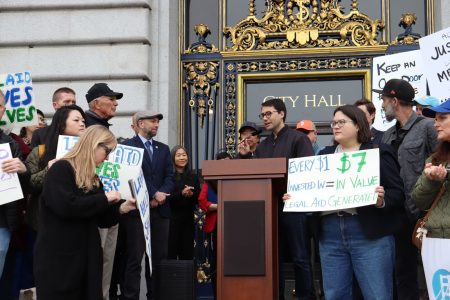 A group of people gathers outside city hall holding signs in support of legal aid, while a speaker addresses the crowd from a podium.